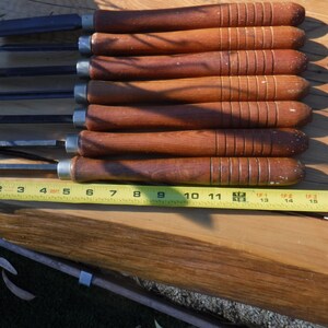May include: Set of eight wood carving tools with brown wooden handles and metal blades. The tools are laid out on a wooden surface with a yellow measuring tape.