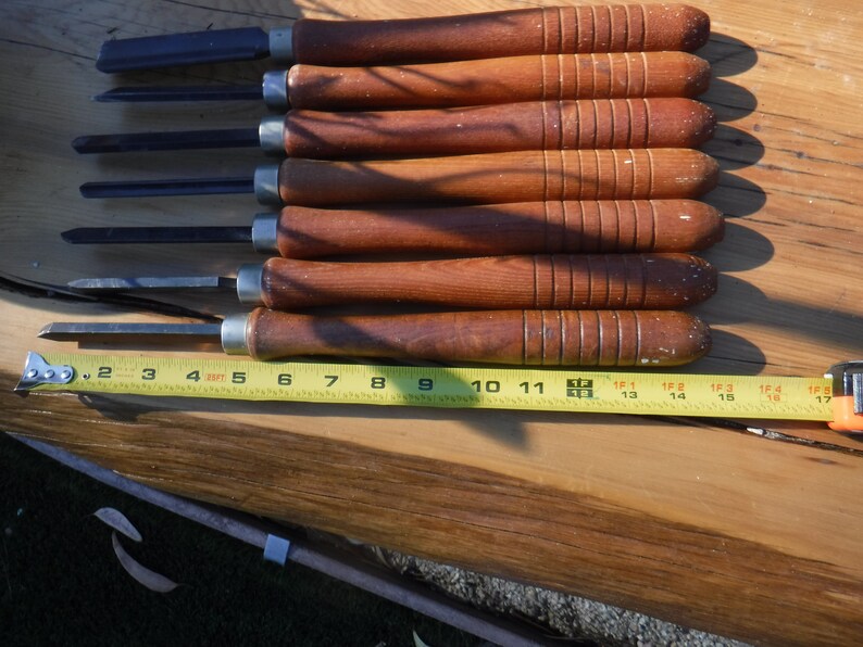 May include: Set of eight wood carving tools with brown wooden handles and metal blades. The tools are laid out on a wooden surface with a yellow measuring tape.
