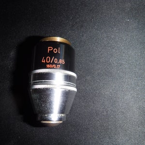May include: A silver microscope objective lens with a black barrel and a gold band. The lens is labeled "Pol 40/0.85 160/0.17".
