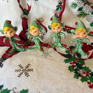 May include: Set of four vintage ceramic Christmas elves. The elves are green with red hats and shoes. They are standing on a white tablecloth with a red and green holiday pattern.