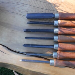 May include: A set of six wood carving tools with wooden handles and metal blades. The tools are arranged in a row on a wooden surface.