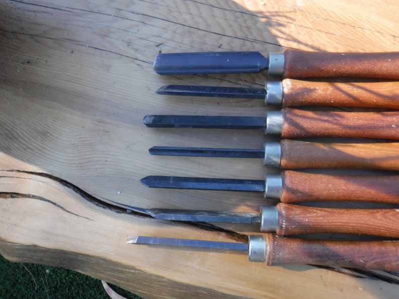 May include: A set of six wood carving tools with wooden handles and metal blades. The tools are arranged in a row on a wooden surface.