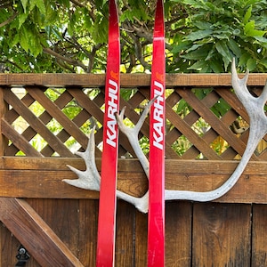 May include: A pair of red cross-country skis with the word "KARHU" printed on them. The skis are leaning against a wooden fence with a white antler.