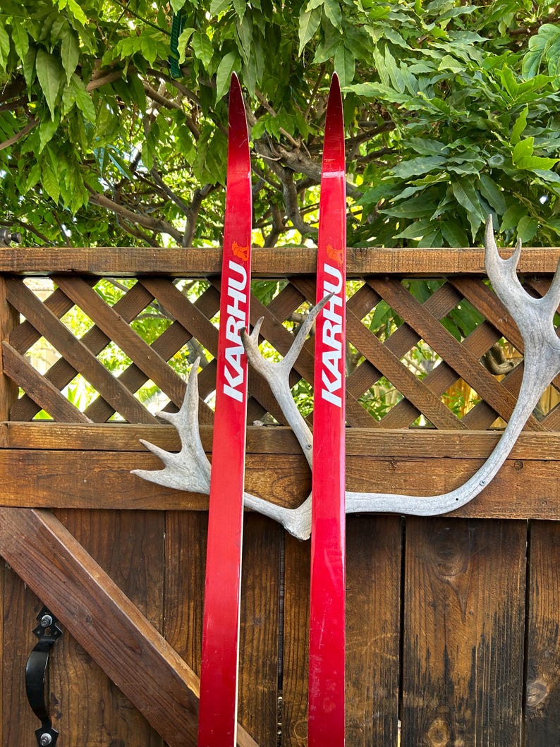 May include: A pair of red cross-country skis with the word "KARHU" printed on them. The skis are leaning against a wooden fence with a white antler.