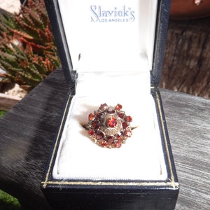 May include: A red gemstone ring with a cluster of small stones set in a gold band. The ring is in a black box with a white interior and the label "Slavick's Los Angeles" on the lid.