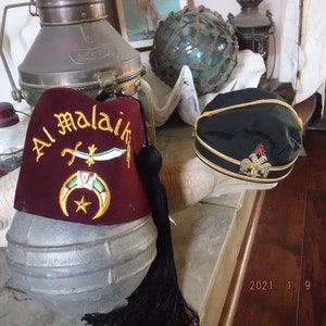 May include: Two fez hats, one red with gold embroidery and the text "Al Malaik" and the other black with gold trim and a gold emblem.
