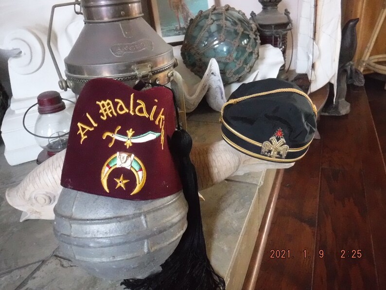 May include: Two fez hats, one red with gold embroidery and the text "Al Malaik" and the other black with gold trim and a gold emblem.