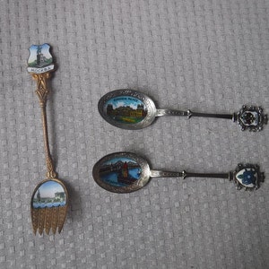 May include: Three silver souvenir spoons with painted images of buildings and the word "MOCKBA" on one spoon. The spoons are on a white textured surface.