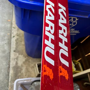 May include: A pair of red skis with the word "KARHU" printed in white on the side. The skis have orange bear logos on each side.