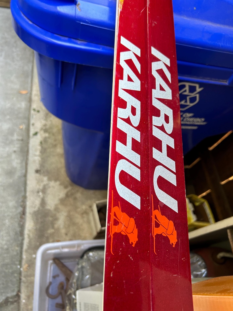 May include: A pair of red skis with the word "KARHU" printed in white on the side. The skis have orange bear logos on each side.