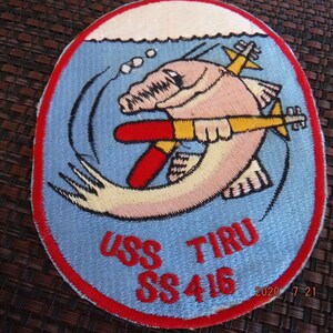 May include: A blue and white embroidered patch with a cartoon fish holding a torpedo. The patch says "USS TIRU SS 416" in red lettering.