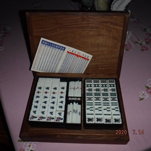 May include: A wooden box containing a set of mahjong tiles. The tiles are white with black and red characters. The box is open and the tiles are arranged in rows. The box has a dark wood finish and is decorated with a simple design.