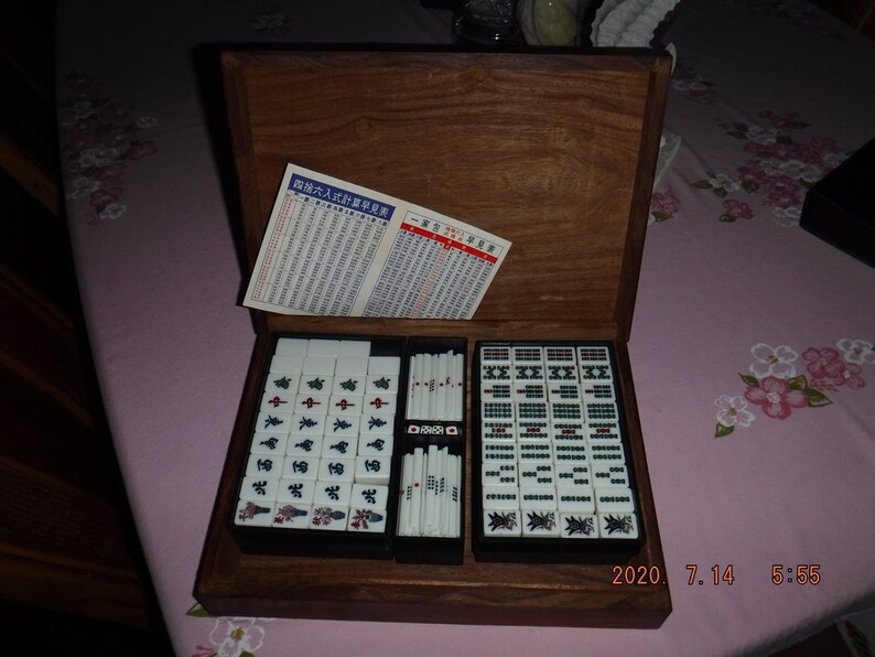 May include: A wooden box containing a set of mahjong tiles. The tiles are white with black and red characters. The box is open and the tiles are arranged in rows. The box has a dark wood finish and is decorated with a simple design.