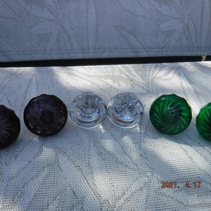 May include: Six glass knobs with a decorative swirl pattern. Two are clear, two are purple, and two are green.