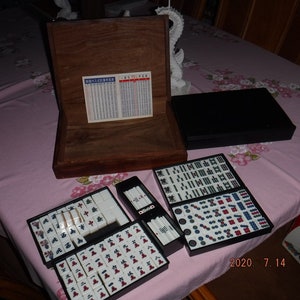 May include: A wooden box with a black lid containing a set of mahjong tiles. The tiles are arranged in a grid pattern and are white with black and red characters. The box also includes a set of dice and a rule book.