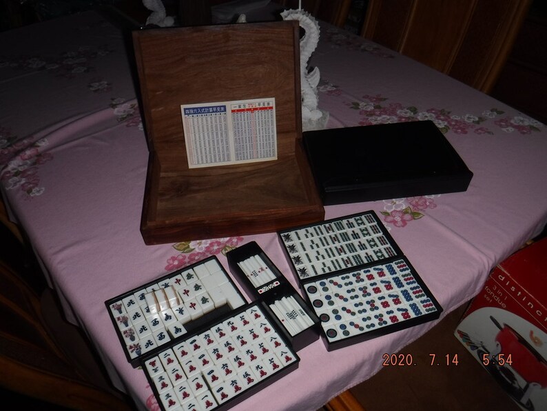 May include: A wooden box with a black lid containing a set of mahjong tiles. The tiles are arranged in a grid pattern and are white with black and red characters. The box also includes a set of dice and a rule book.