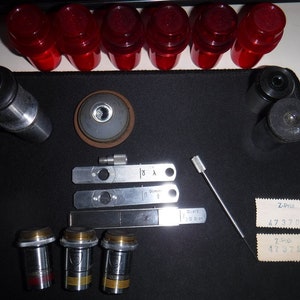 May include: A collection of scientific equipment and components, including red lenses, cylindrical metal pieces, and small rectangular metal plates. The items are arranged on a black surface, with two labels visible.