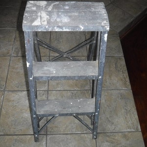 May include: A two-step metal step stool with wooden steps. The steps are painted black and have white paint splatters. The stool is standing on a tiled floor.