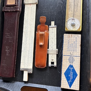 May include: A collection of vintage slide rules and a protractor, displayed on a dark tray. The items include a large slide rule, a smaller one in a leather case, and a protractor in a yellow box. The text "tractograt" is visible.