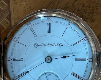 May include: A gold-toned Elgin Natl. Watch Co. pocket watch with a white face and black hands. The watch features Roman numerals and a smaller seconds dial. The watch is open, revealing the face and the intricate details of the timepiece.