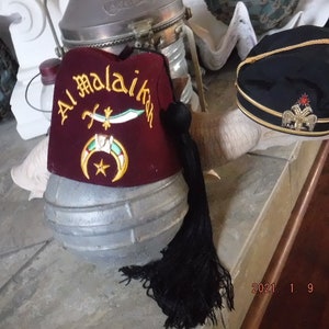 May include: Two red and black fez hats with gold embroidery. The fez on the left has the text "Al Malaikah" embroidered on it. The fez on the right has a gold eagle emblem with a red star.