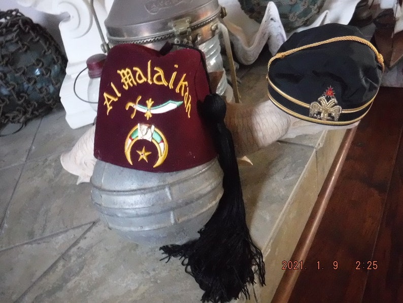 May include: Two red and black fez hats with gold embroidery. The fez on the left has the text "Al Malaikah" embroidered on it. The fez on the right has a gold eagle emblem with a red star.