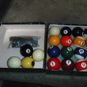 May include: A set of 13 billiard balls in a black and white case. The balls are numbered 1 through 13 and are white, red, yellow, and green. The case is open and the balls are arranged in a circle.