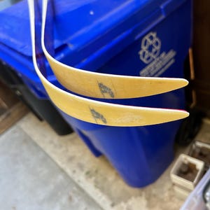 May include: Two wooden recurve bow limbs with white tips and red accents. The limbs are curved and have a slight yellow tint. The limbs are resting on top of a blue recycling bin.