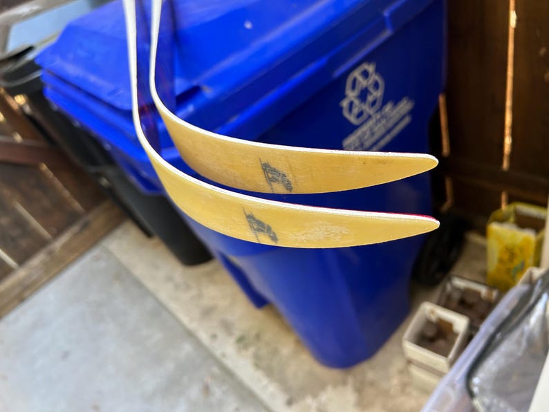 May include: Two wooden recurve bow limbs with white tips and red accents. The limbs are curved and have a slight yellow tint. The limbs are resting on top of a blue recycling bin.