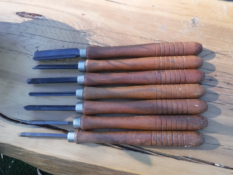 May include: A set of seven wood carving tools with wooden handles. The tools have different blade shapes and sizes.