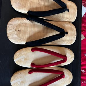 May include: Four pairs of traditional Japanese geta sandals. Two pairs have natural-colored wooden soles with black fabric straps. The other two pairs have natural-colored wooden soles with red fabric straps. The sandals are displayed on a black tray.