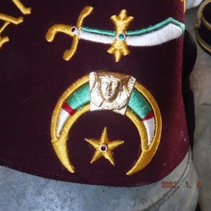 May include: A maroon fez with gold embroidery of a crescent moon, a five-pointed star, and a face in a crown. The fez also has a white and green embroidered design.