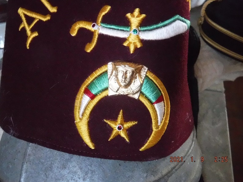 May include: A maroon fez with gold embroidery of a crescent moon, a five-pointed star, and a face in a crown. The fez also has a white and green embroidered design.