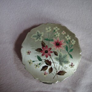 May include: A silver compact with a floral design. The flowers are pink, red, and white with green leaves. The compact has a scalloped edge.