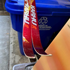 May include: Two red and white ski poles with the word "KARHU" printed on them. The poles are leaning against a blue recycling bin.