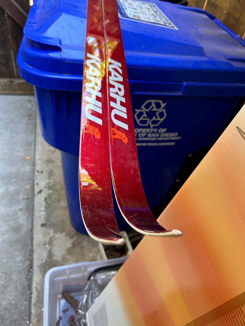 May include: Two red and white ski poles with the word "KARHU" printed on them. The poles are leaning against a blue recycling bin.