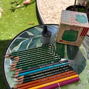 May include: A collection of pencils, including A.W. Faber-Castell 9000, Eagle Writing, and Pedigree Drawing pencils, arranged on a mosaic table. A pencil sharpener and a vintage Vari-Point box are also present. The pencils vary in color and lead type.