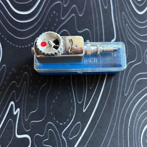 May include: A silver-toned mechanical timer with a circular dial featuring a red indicator dot and a black arrow. The device has a metallic body, a knurled adjustment knob, and a protruding metal tip, resting on a rectangular blue plastic case.