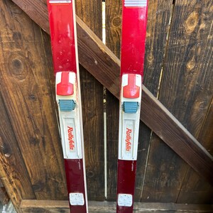 May include: A pair of red vintage cross-country skis with white bindings and the text "Rollefella" printed on the side.