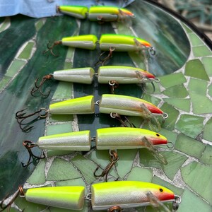 May include: A collection of fishing lures arranged on a mosaic table. The lures are primarily white, yellow, and red, with attached hooks. The lures are designed to mimic small fish, with a segmented body and a propeller-like tail.