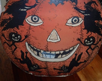 May include: A vintage Halloween decoration featuring a large orange pumpkin with a wide, toothy grin. The pumpkin has black eyes and a triangular nose. Black silhouettes of witches and cats surround the pumpkin.