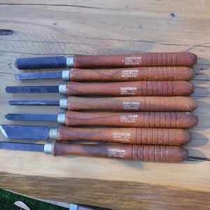 May include: A set of eight wood-handled carving tools with black metal blades. The tools are labeled "CRAFTSMAN HIGH SPEED STEEL" and are arranged in a row on a wooden surface.