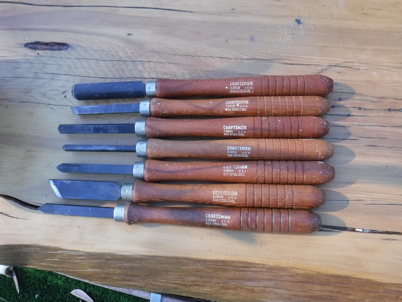 May include: A set of eight wood-handled carving tools with black metal blades. The tools are labeled "CRAFTSMAN HIGH SPEED STEEL" and are arranged in a row on a wooden surface.