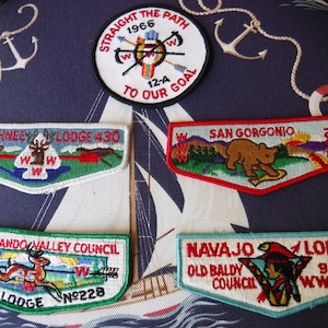 May include: Four embroidered patches with a scout theme. The top patch is a compass with the text "Straight the Path 1966 12-A To Our Goal". The other three patches are rectangular and feature a lodge name, location, and number. The patches are on a blue and white striped fabric background.