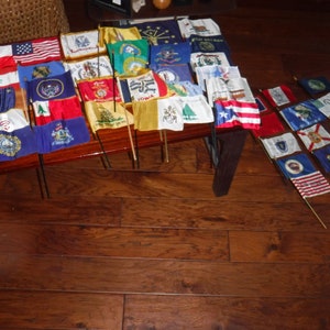May include: A collection of small state flags displayed on wooden sticks. The flags are arranged in rows on a wooden table and on the floor. The flags are all different colors and designs, representing different states in the United States.