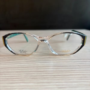 Può includere: Occhiali trasparenti con forma a occhi di gatto. Le montature presentano dettagli neri, dorati e turchese. Il testo "Collection ELITE" e "DAMY" è stampato sulle lenti.