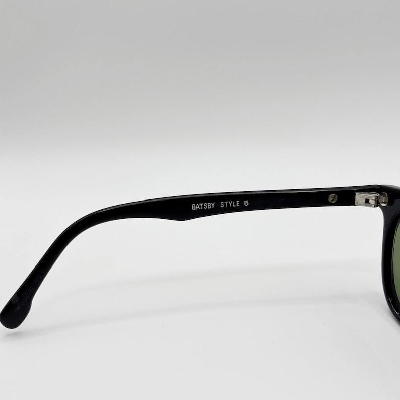 May include: Black sunglasses with the text "Gatsby Style 5" on the arm. The glasses have a classic design with a curved frame and dark lenses. The image is set against a plain white background.