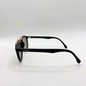 May include: Black sunglasses with a gold-colored top bar and dark green lenses. The sunglasses have a classic design with a rectangular frame and curved temples. The brand logo is visible on the temple.