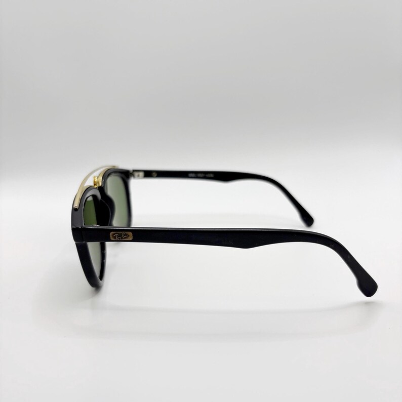 May include: Black sunglasses with a gold-colored top bar and dark green lenses. The sunglasses have a classic design with a rectangular frame and curved temples. The brand logo is visible on the temple.