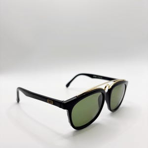 May include: Black sunglasses with green lenses and a gold-colored metal bridge. The sunglasses have a classic design with a rectangular frame and a subtle logo on the temple. The lenses are a dark green color.